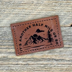 May include: Brown leather patch with black stitching and the text "It's another half mile or so" with a mountain and tree graphic.