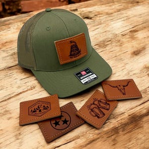 Può includere: Un cappello da camionista verde con una toppa in pelle marrone che dice "Don't Tread on Me" e un serpente. Ci sono cinque toppe in pelle marrone aggiuntive con vari disegni, tra cui un teschio, montagne, boccali di birra e una stella.