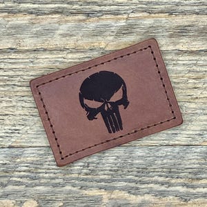 May include: A rectangular, brown leather patch with a black skull graphic. The patch is stitched around the edges and has a distressed appearance. The skull graphic is a stylized representation of a human skull.
