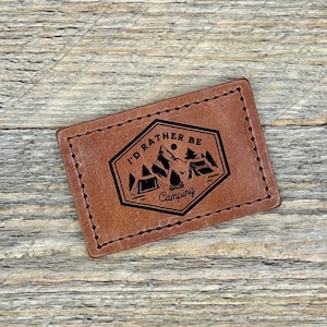 Peut inclure: Un patch en cuir marron avec un graphique noir et blanc d'une chaîne de montagnes, deux tentes et une lune. Le texte "I'd rather be Camping" est imprimé en noir.