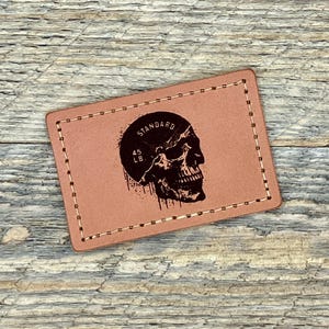 Puede incluir: Un parche rectangular de cuero color canela con un borde cosido. Un gráfico de calavera negra está centrado, dentro de un círculo negro, con la palabra "STANDARD" arriba y "45 LB." abajo. El parche está sobre una superficie de madera.