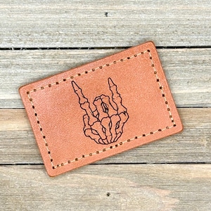 Pode incluir: Um remendo de couro marrom com uma borda costurada. O remendo apresenta uma mão de esqueleto em preto e branco fazendo o gesto de rock and roll.