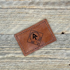 May include: A rectangular brown leather patch with black stitching around the edges. The patch features the words "Appalachian Trail" in a diamond shape, with an "A" and an arrow pointing upwards. Below the diamond are the words "Katahdin Maine to Springer Mtn Georgia 2,189 Miles."