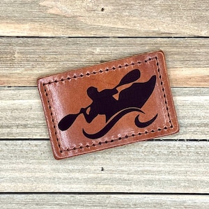 May include: A brown leather keychain with a black silhouette of a person kayaking. The keychain has a stitched edge.