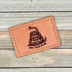 May include: A brown leather patch with a stitched border. The patch features a coiled rattlesnake with the text "Don't Tread On Me".