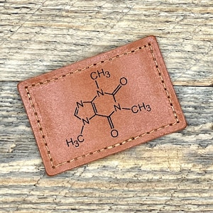 May include: A brown leather keychain with a chemical formula for caffeine printed on it. The formula is surrounded by a stitched border.