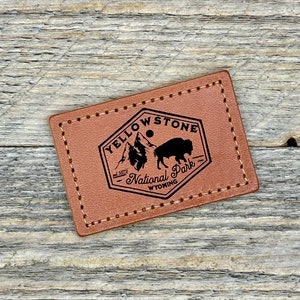 May include: A brown leather patch with a black outline and stitching. The patch features a black silhouette of a bison and mountains with the text "Yellowstone National Park, est. 1872, Wyoming".