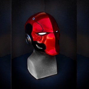 May include: A glossy red helmet with black accents, resembling a superhero or comic book character. The helmet features white eye slits and a metallic silver earpiece. It is displayed on a gray mannequin head against a dark background.