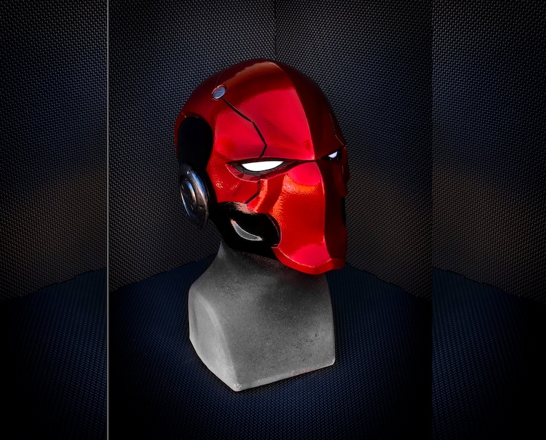 May include: A glossy red helmet with black accents, resembling a superhero or comic book character. The helmet features white eye slits and a metallic silver earpiece. It is displayed on a gray mannequin head against a dark background.