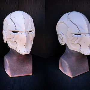 May include: A white, futuristic-style helmet with a segmented design. The helmet features angular lines, eye openings, and ear details. It is displayed on a brown mannequin head against a dark background. The helmet has a textured surface with dark gray accent lines.