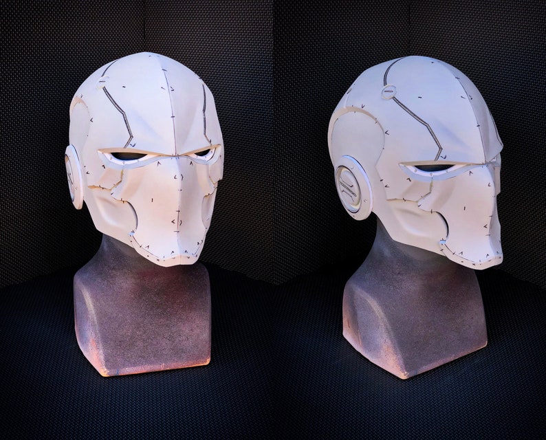 May include: A white, futuristic-style helmet with a segmented design. The helmet features angular lines, eye openings, and ear details. It is displayed on a brown mannequin head against a dark background. The helmet has a textured surface with dark gray accent lines.