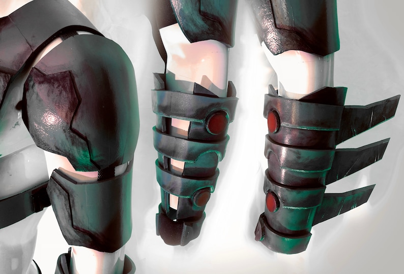 May include: Three close-up views of a dark grey and green metal forearm armour piece with red accents. The armour has a segmented design with a series of plates and a pointed edge.