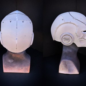 May include: A white, futuristic-style helmet with a detailed design, featuring visible seams and rivets. The helmet is displayed on a mannequin head, showcasing its shape and form. The design includes a circular emblem on the side and a visor area.