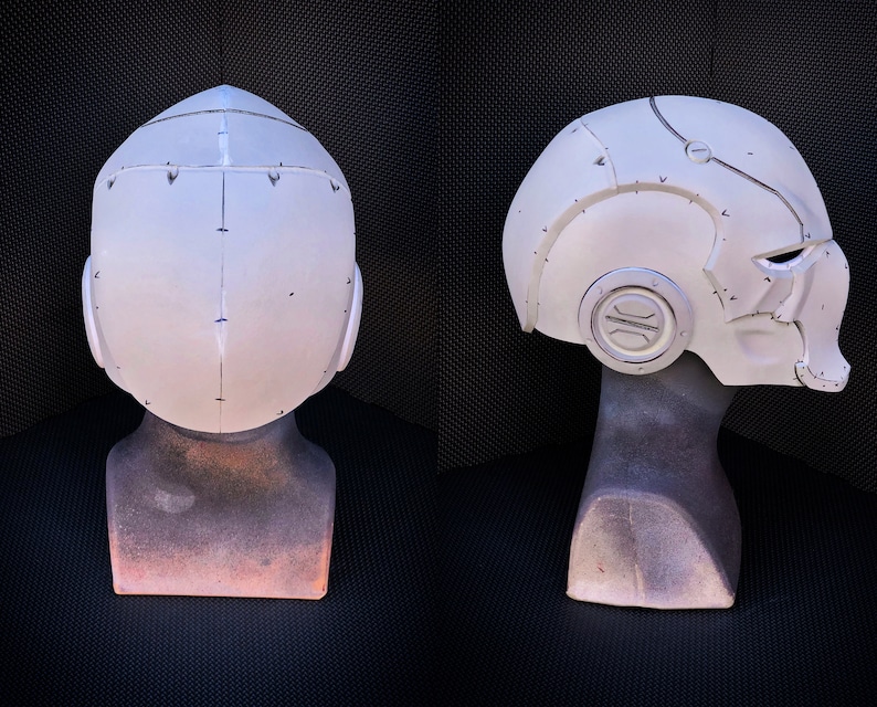 May include: A white, futuristic-style helmet with a detailed design, featuring visible seams and rivets. The helmet is displayed on a mannequin head, showcasing its shape and form. The design includes a circular emblem on the side and a visor area.