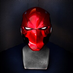May include: A glossy red helmet with white glowing eye slits, displayed on a gray stand. The helmet has a distinctive angular design and a smooth, reflective surface. The background is a dark, textured fabric.