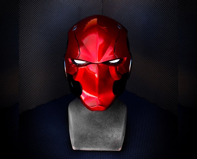 May include: A glossy red helmet with white glowing eye slits, displayed on a gray stand. The helmet has a distinctive angular design and a smooth, reflective surface. The background is a dark, textured fabric.