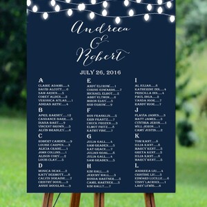 May include: A navy blue wedding seating chart with white lights strung across the top. The chart lists the names of guests in alphabetical order with their table assignments. The text at the top reads "Andreea & Robert" and "July 26, 2016".