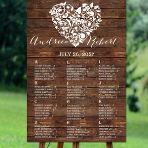May include: A rustic wooden wedding seating chart with a white heart design and the names Andreea and Robert. The chart lists guest names alphabetically with table numbers. The date July 26, 202? is printed at the top.