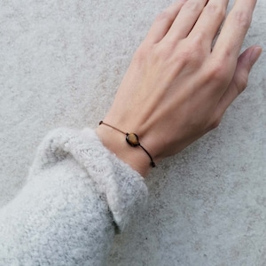 Puede incluir: Un brazalete de cuerda marrón con una sola cuenta de piedra preciosa marrón y dorada.