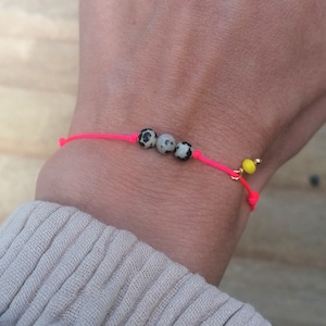 Puede incluir: Un brazalete de cuerda rosa con tres cuentas moteadas de negro y blanco y un dije de cuenta amarilla.