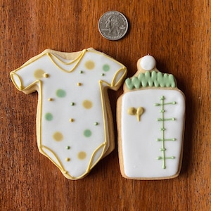 May include: Two decorated sugar cookies, one shaped like a baby onesie with green and gold polka dots, and the other shaped like a baby bottle with green and white details.