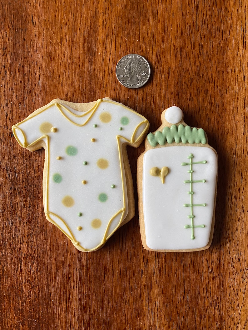 May include: Two decorated sugar cookies, one shaped like a baby onesie with green and gold polka dots, and the other shaped like a baby bottle with green and white details.