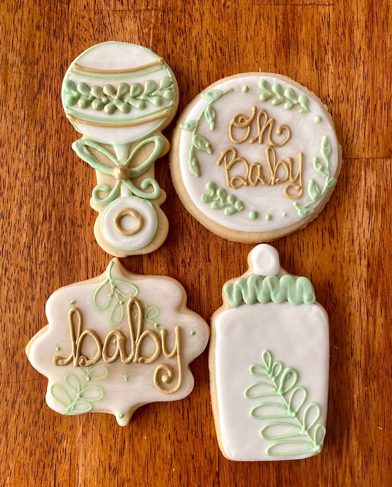Pu&ograve; includere: Quattro biscotti di zucchero decorati con glassa verde e oro. I biscotti hanno la forma di un sonaglio, un biberon, un cerchio e uno stendardo. Il biscotto rotondo ha le parole "Oh Baby" scritte su di esso. Il biscotto a forma di stendardo ha la parola "baby" scritta su di esso.