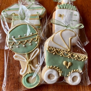 May include: Four decorated sugar cookies in individual cellophane bags. The cookies are decorated with green and gold icing and feature a baby bottle, a rattle, a onesie, and a baby carriage.