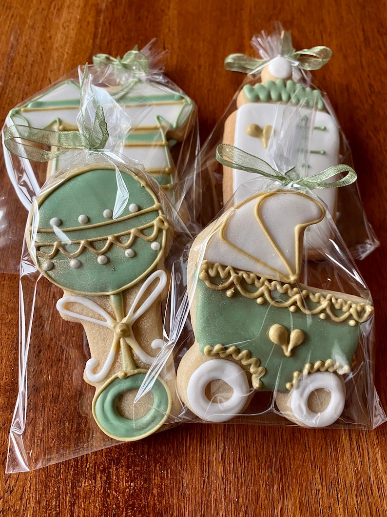 May include: Four decorated sugar cookies in individual cellophane bags. The cookies are decorated with green and gold icing and feature a baby bottle, a rattle, a onesie, and a baby carriage.