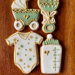 May include: Four decorated sugar cookies in a baby shower theme. The cookies are a baby carriage, a rattle, a baby onesie, and a baby bottle. The cookies are decorated with green and gold icing.