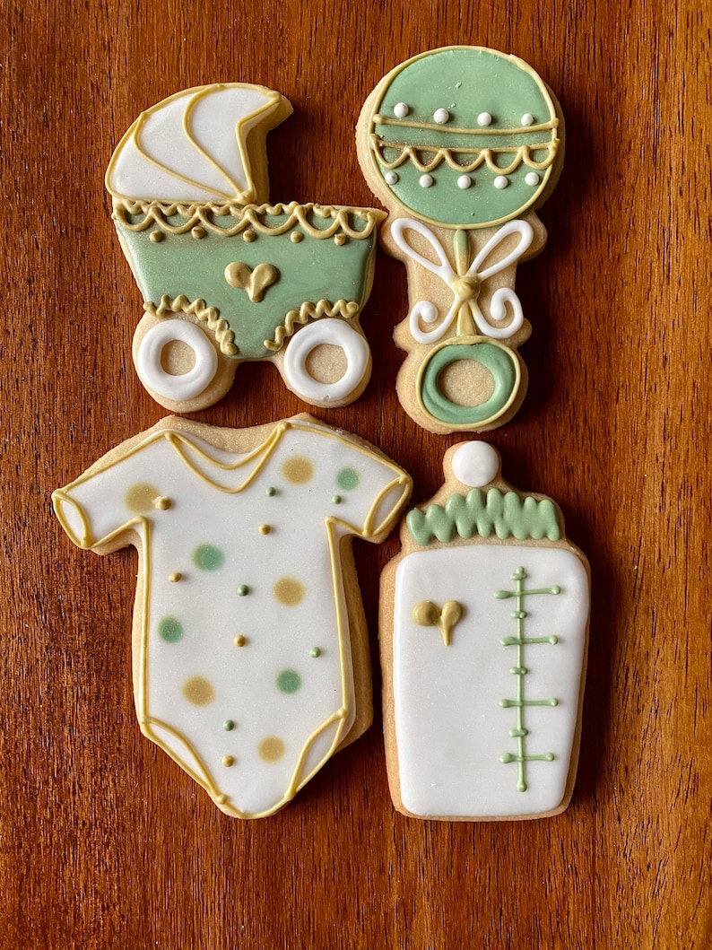 May include: Four decorated sugar cookies in a baby shower theme. The cookies are a baby carriage, a rattle, a baby onesie, and a baby bottle. The cookies are decorated with green and gold icing.