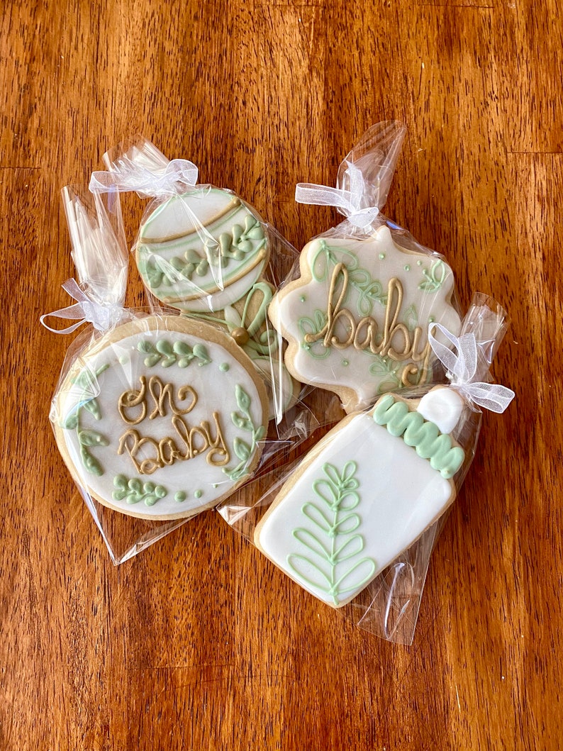 Pu&ograve; includere: Quattro biscotti di zucchero decorati in sacchetti di cellophane individuali. I biscotti sono decorati con glassa verde e oro. I biscotti mostrano le parole "One Baby", "Baby", uno sonaglio e un biberon.