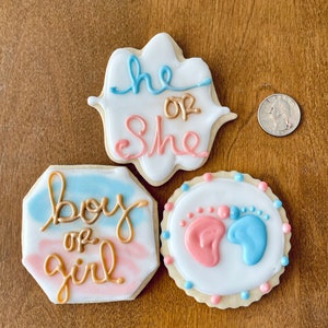 Puede incluir: Tres galletas de az&uacute;car decoradas. Una galleta es blanca con glaseado azul y dorado y dice "He or She". Otra galleta es azul y rosa con glaseado dorado y dice "Boy or Girl". La &uacute;ltima galleta es blanca con glaseado azul y rosa y tiene dos huellas de pies, una rosa y una azul.