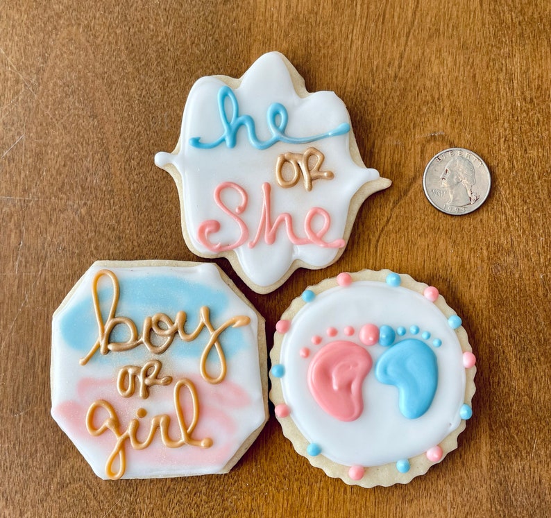 Puede incluir: Tres galletas de az&uacute;car decoradas. Una galleta es blanca con glaseado azul y dorado y dice "He or She". Otra galleta es azul y rosa con glaseado dorado y dice "Boy or Girl". La &uacute;ltima galleta es blanca con glaseado azul y rosa y tiene dos huellas de pies, una rosa y una azul.