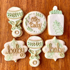 Pu&ograve; includere: Sei biscotti di zucchero decorati con glassa verde e oro. I biscotti raffigurano un biberon, un sonaglio e le parole "Oh Baby" e "Baby".