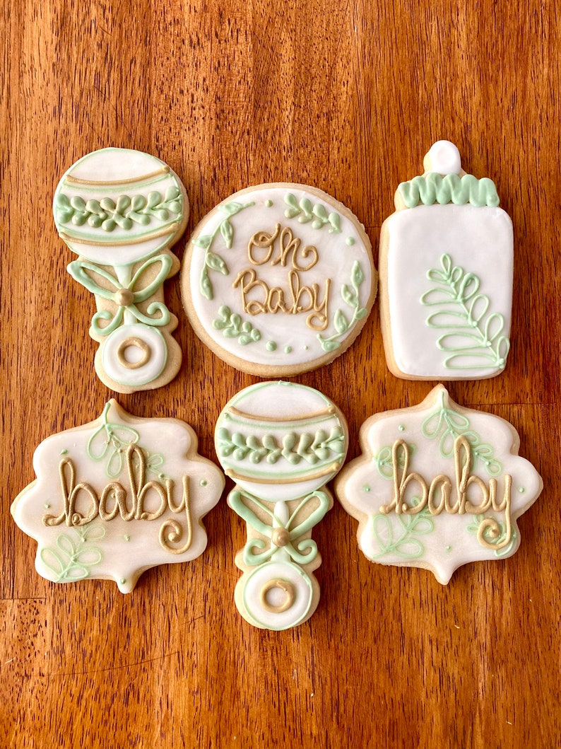 Pu&ograve; includere: Sei biscotti di zucchero decorati con glassa verde e oro. I biscotti raffigurano un biberon, un sonaglio e le parole "Oh Baby" e "Baby".