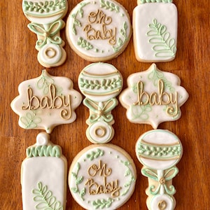 Pu&ograve; includere: Nove biscotti di zucchero decorati con glassa verde e oro. I biscotti presentano immagini di biberon, sonagli e il testo "Oh Baby" e "Baby".
