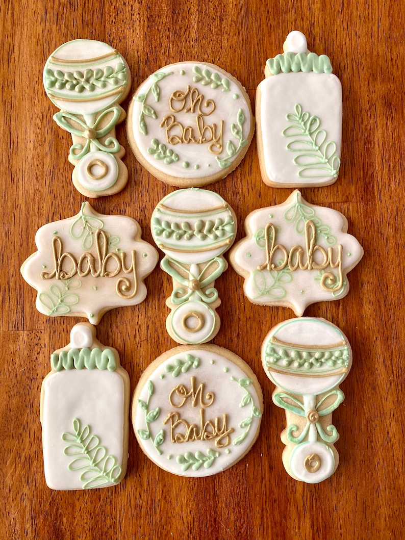 Pu&ograve; includere: Nove biscotti di zucchero decorati con glassa verde e oro. I biscotti presentano immagini di biberon, sonagli e il testo "Oh Baby" e "Baby".