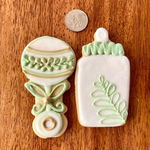 Pu&ograve; includere: Due biscotti di zucchero decorati, uno a forma di biberon e l'altro a forma di sonaglio. Entrambi i biscotti sono bianchi con glassa verde e dorata.