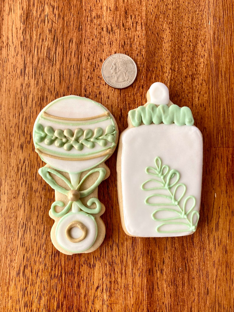 Pu&ograve; includere: Due biscotti di zucchero decorati, uno a forma di biberon e l'altro a forma di sonaglio. Entrambi i biscotti sono bianchi con glassa verde e dorata.