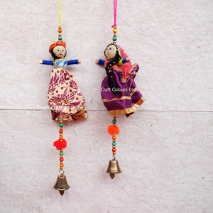May include: Two handmade fabric dolls hanging from strings. One doll wears a turban and patterned skirt, the other a headscarf and purple skirt. Both have beaded accents, orange pom-poms, and brass bells. The text "Craft Colours India" is visible.