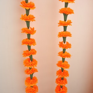 Puede incluir: Dos hileras verticales de guirnaldas de cal&eacute;ndulas de color naranja brillante sobre un fondo blanco. Cada flor es de un naranja vibrante, con tallos verdes. Estas guirnaldas se utilizan probablemente con fines decorativos.