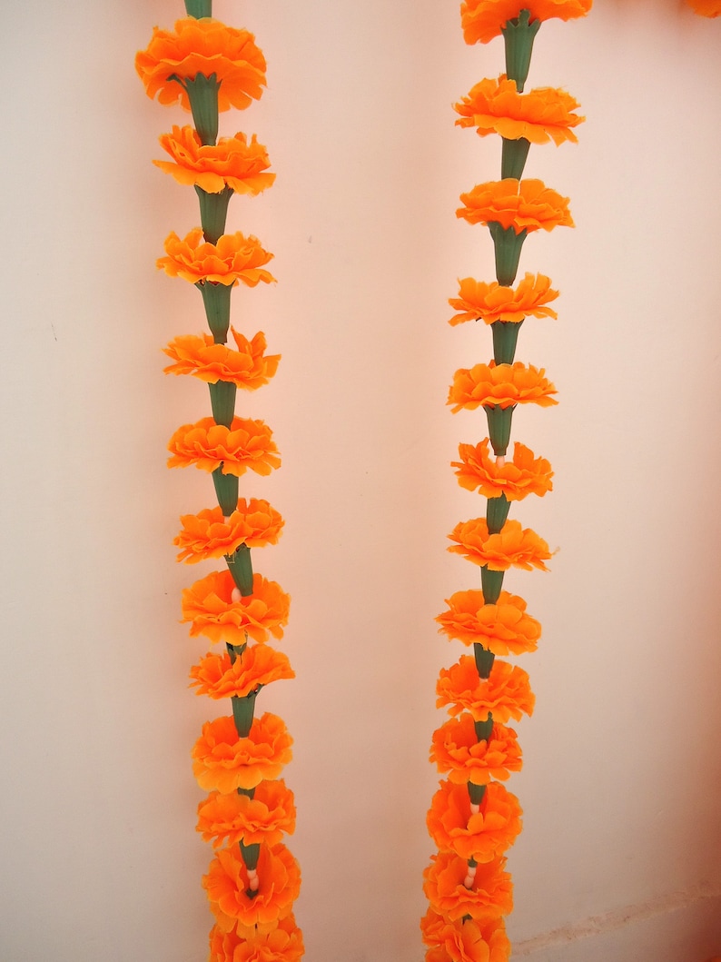 Puede incluir: Dos hileras verticales de guirnaldas de cal&eacute;ndulas de color naranja brillante sobre un fondo blanco. Cada flor es de un naranja vibrante, con tallos verdes. Estas guirnaldas se utilizan probablemente con fines decorativos.