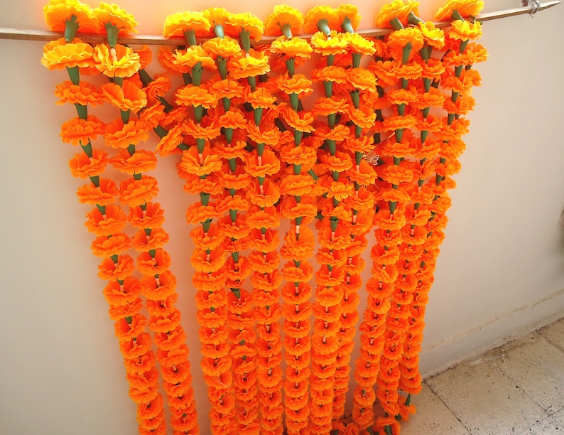 Puede incluir: Una colecci&oacute;n de guirnaldas de flores de cal&eacute;ndula de color naranja vibrante, cada una ensartada con tallos verdes. Las guirnaldas est&aacute;n suspendidas de una vara de madera, creando una exhibici&oacute;n decorativa. Las flores est&aacute;n en plena floraci&oacute;n, mostrando sus p&eacute;talos en capas.
