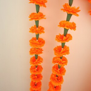 Puede incluir: Primer plano de dos guirnaldas de flores de cal&eacute;ndula de color naranja brillante, cada flor conectada por un tallo verde. Las cal&eacute;ndulas est&aacute;n en plena floraci&oacute;n, con m&uacute;ltiples capas de p&eacute;talos. El fondo es de color blanco roto liso.