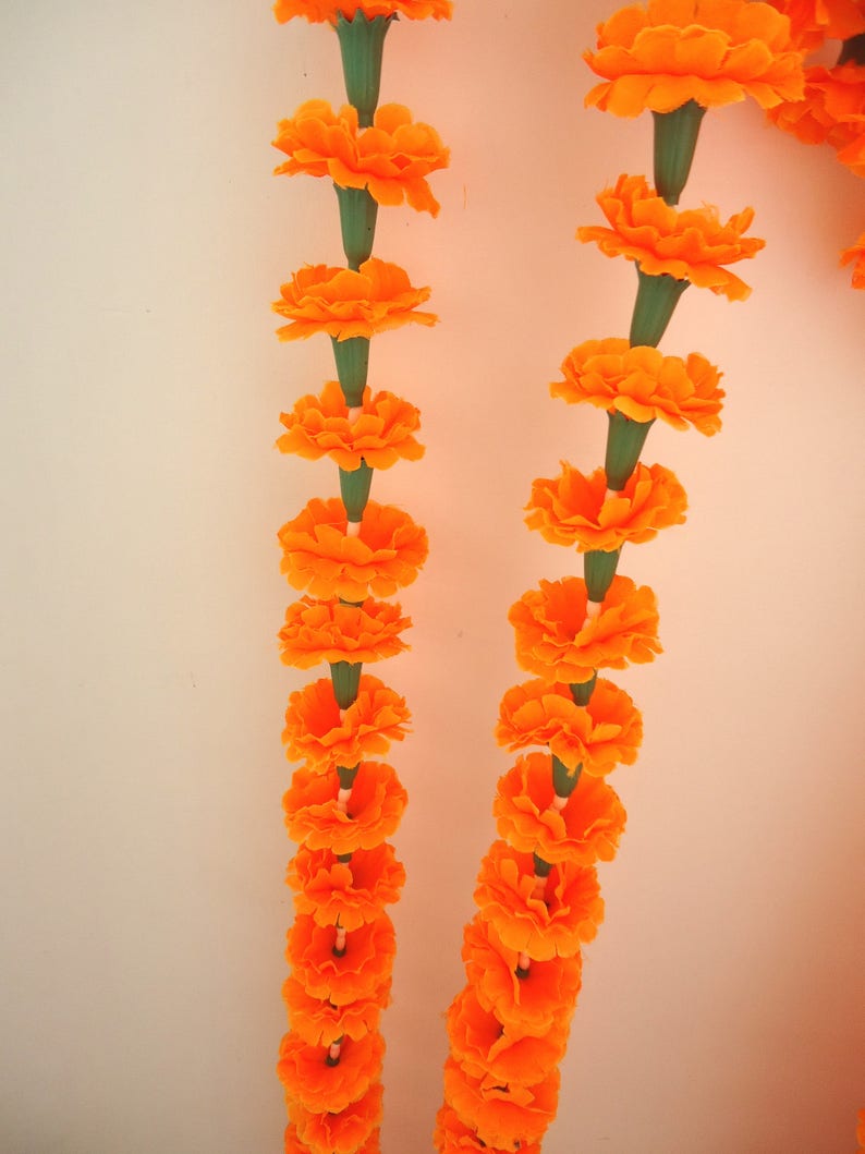 Puede incluir: Primer plano de dos guirnaldas de flores de cal&eacute;ndula de color naranja brillante, cada flor conectada por un tallo verde. Las cal&eacute;ndulas est&aacute;n en plena floraci&oacute;n, con m&uacute;ltiples capas de p&eacute;talos. El fondo es de color blanco roto liso.