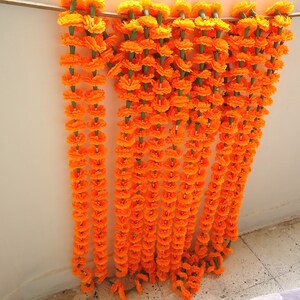 Puede incluir: Varias guirnaldas de flores de cal&eacute;ndula de color naranja brillante, con tallos verdes, colgadas de una barra de madera. Las guirnaldas son una exhibici&oacute;n vibrante, ideal para celebraciones y decoraciones, agregando un toque de color.