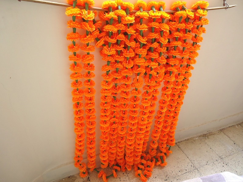 Puede incluir: Varias guirnaldas de flores de cal&eacute;ndula de color naranja brillante, con tallos verdes, colgadas de una barra de madera. Las guirnaldas son una exhibici&oacute;n vibrante, ideal para celebraciones y decoraciones, agregando un toque de color.