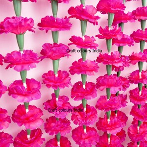 May include: Rows of vibrant fuchsia artificial marigold flowers, each with a green stem, hang against a light pink background. The flowers are densely packed, creating a visually striking display. The text "Craft colours India" is visible.