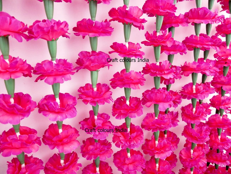 May include: Rows of vibrant fuchsia artificial marigold flowers, each with a green stem, hang against a light pink background. The flowers are densely packed, creating a visually striking display. The text "Craft colours India" is visible.